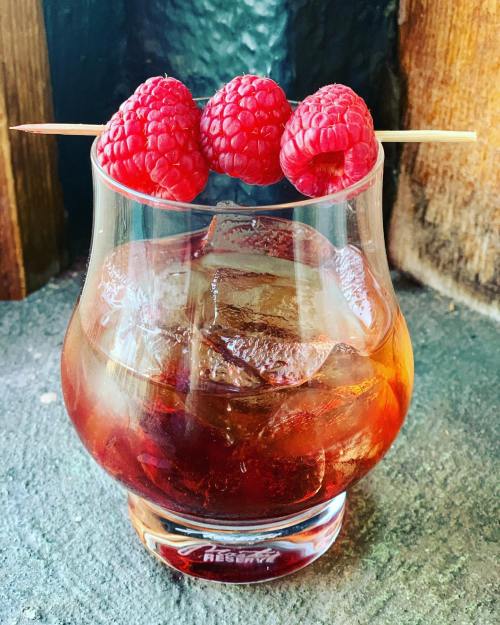 A glass of iced drink garnished with three fresh raspberries on a skewer, set against a rustic background.