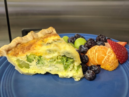 A slice of broccoli quiche on a blue plate, accompanied by grapes, blueberries, strawberries, and orange segments.