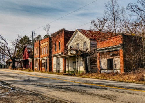 Abandoned buildings line a quiet road, with overgrown vegetation and a cloudy sky overhead.