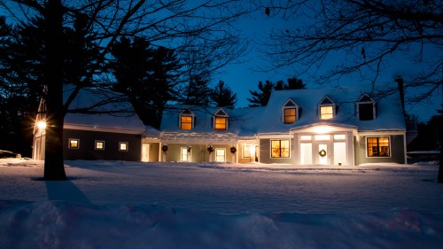 A cozy house at night, surrounded by snow, with warm lights glowing from the windows and a wreath on the door.