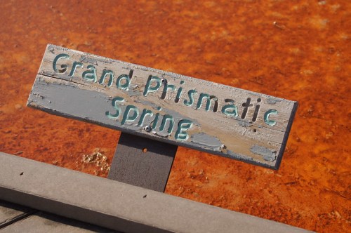 Sign reading "Grand Prismatic Spring" in front of a vibrant orange and blue geothermal spring.
