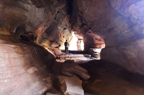 A person stands inside a colorful cave, surrounded by textured rock formations and natural light filtering through openings.