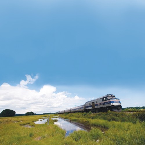 A train travels through a lush green landscape under a blue sky with scattered clouds, reflecting in a nearby water body.