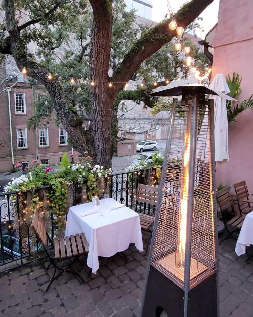 Cozy outdoor dining area with string lights, a patio heater, and tables set for two, surrounded by greenery.