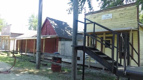 Old western-style buildings with wooden stairs and a sign reading "Gun Fighters." Trees and a dirt path are visible.