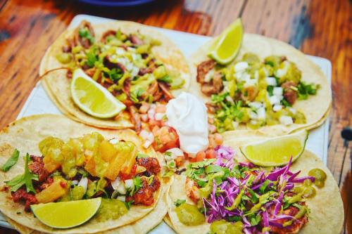 A platter of four tacos topped with various ingredients, lime wedges, and a dollop of sour cream on a wooden table.