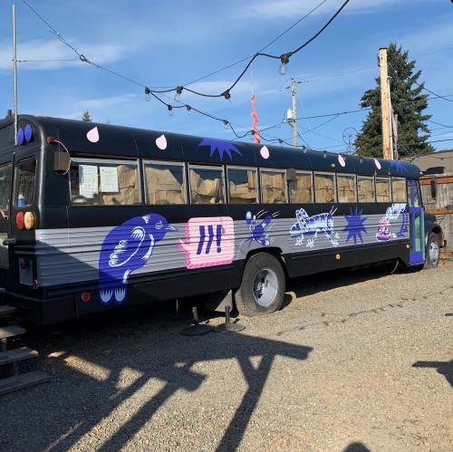 A colorful, artistic bus with various designs parked in a gravel area under a clear blue sky.
