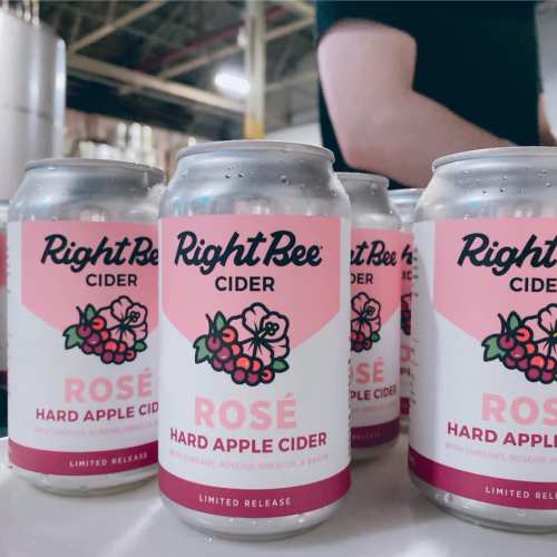 Cans of Right Bee Cider Rosé hard apple cider on a table, with a person’s hand in the background.