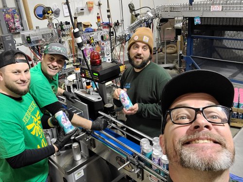 Four men in a brewery are smiling and working on a canning line, holding colorful cans in a lively production environment.