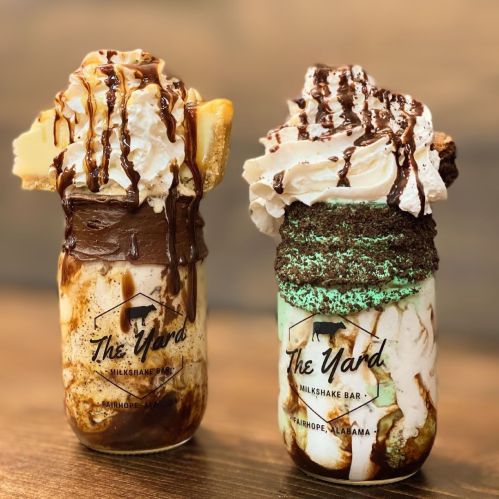 Two decadent milkshakes in jars, topped with whipped cream, chocolate drizzle, and various toppings, from The Yard Milkshake Bar.