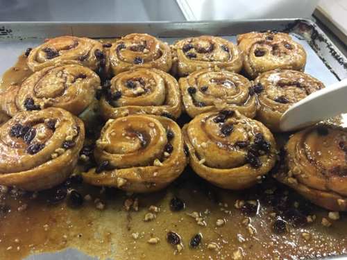 Freshly baked cinnamon rolls with raisins and nuts, glistening with syrup on a baking tray.