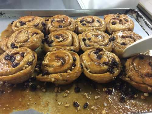 Freshly baked cinnamon rolls with raisins and nuts, glistening with syrup on a baking tray.