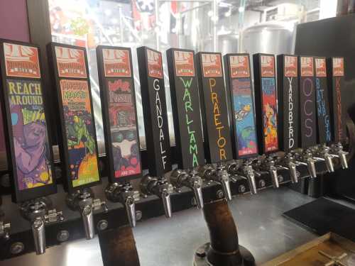 A row of colorful beer taps with unique labels, showcasing various craft beers in a brewery setting.