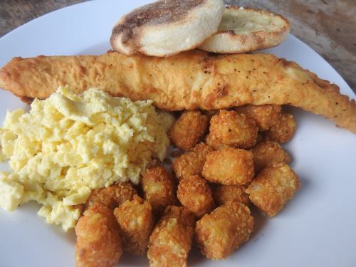 A plate with a fried fish fillet, scrambled eggs, tater tots, and two English muffins.