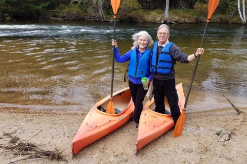 Two people in orange kayaks on a sandy shore, smiling and holding paddles by a river surrounded by trees.