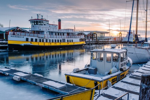 A yellow and white ferry docked at a frozen harbor during sunset, with snow-covered boats and a calm water surface.