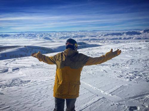 A person in a yellow jacket stands on a snowy mountain, arms outstretched, overlooking a vast winter landscape.