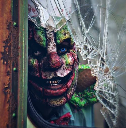 A creepy clown with a painted face and a sinister grin peers through a broken window, adding to the eerie atmosphere.
