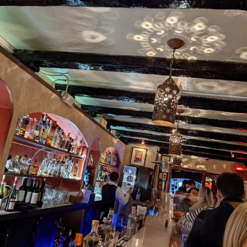 A cozy bar interior with wooden beams, decorative lighting, and shelves filled with various bottles.
