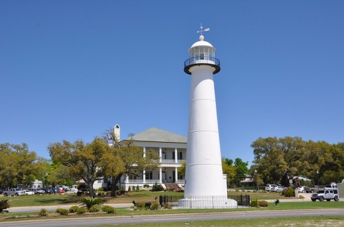 A tall white lighthouse stands beside a historic building, surrounded by greenery and clear blue skies.