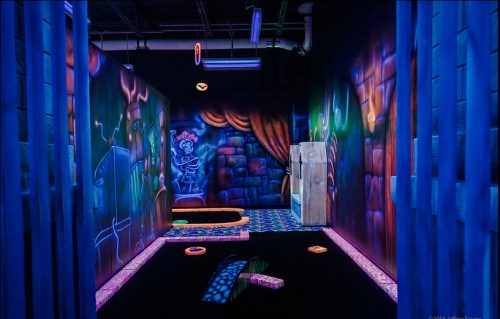 A vibrant, neon-lit mini-golf course with glowing walls, a fridge, and colorful obstacles in a dark room.