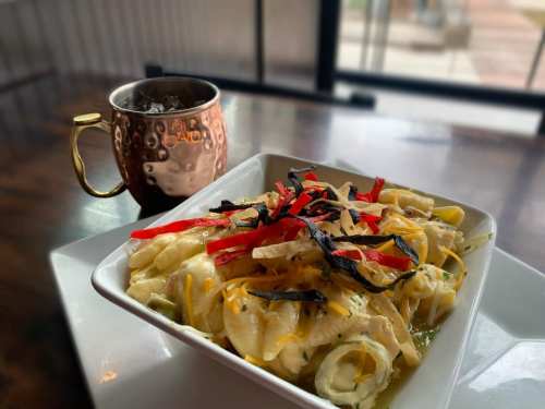 A bowl of creamy pasta topped with red peppers and black olives, alongside a copper mug filled with a dark beverage.
