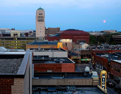 A city skyline at dusk featuring a tall tower, buildings, and a rising moon in the background.