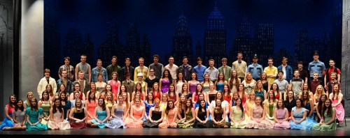 A large group of performers in colorful costumes poses on stage, with a city skyline backdrop.