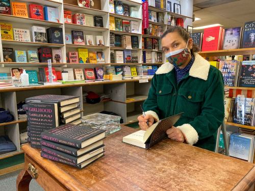 An author signs books at a table in a cozy bookstore filled with shelves of various titles.