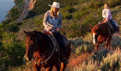 Two women riding horses on a scenic hillside during sunset, surrounded by greenery and a view of the water.