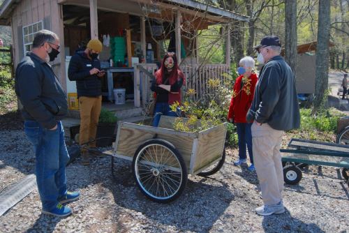 A group of people gathers around a wooden cart outside a building, discussing plants in a natural setting.