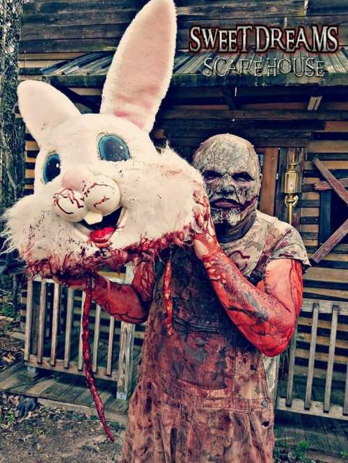 A bloodied figure in a creepy outfit holds a large, cartoonish bunny head outside a haunted house.