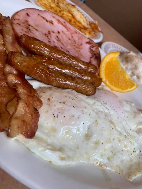 A breakfast plate featuring fried eggs, bacon, sausage links, ham, and a slice of orange.