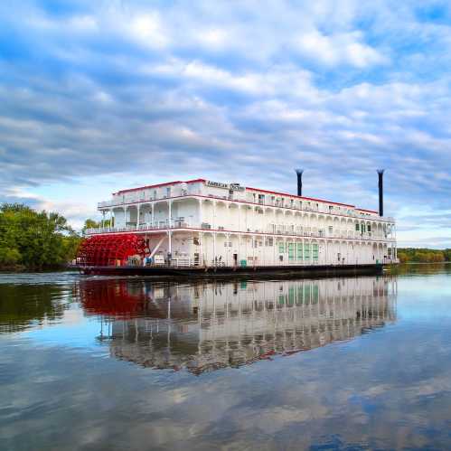 A large riverboat with a red paddle wheel floats on calm water, reflecting clouds and trees in the background.