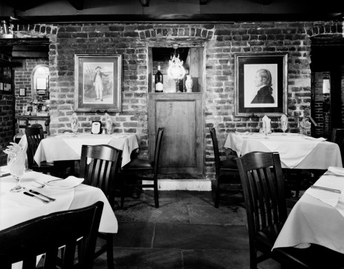 A cozy restaurant interior with wooden tables, white tablecloths, and framed portraits on a brick wall.