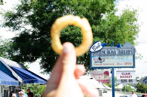 A hand holds up an onion ring in front of Bob's Clam Hut sign, featuring fresh seafood and a blue sky.