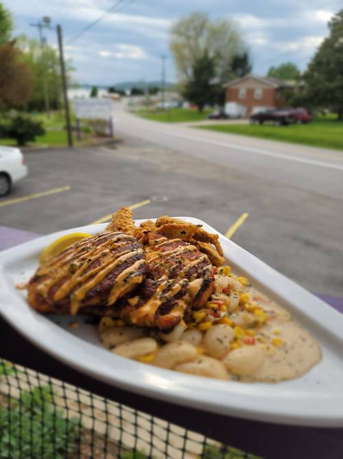 A plate of grilled chicken with a drizzle of sauce, served with corn and creamy sauce, set against a scenic outdoor background.