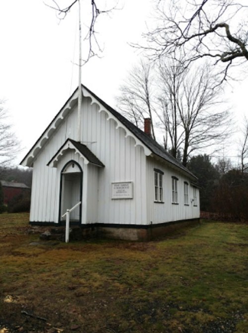A small, white wooden building with a peaked roof, surrounded by bare trees and grass on a cloudy day.