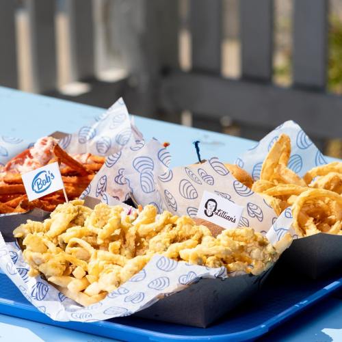 A colorful spread of fried foods, including curly fries, onion rings, and macaroni and cheese, served in paper trays.