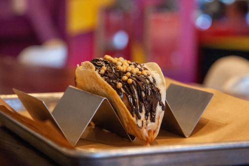 A dessert taco filled with chocolate and topped with crunchy sprinkles, served on a metal tray.