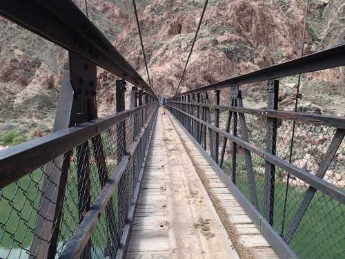 A narrow metal bridge stretches over a river, flanked by rocky cliffs and greenery in the background.