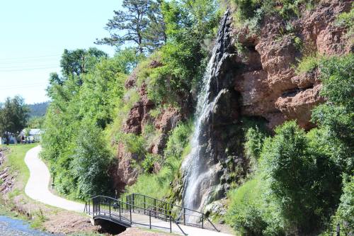 A waterfall cascades down a rocky cliff, surrounded by lush greenery, with a path and bridge nearby.