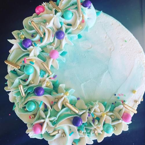 A round cake decorated with teal frosting, colorful sprinkles, and floral designs around the edge.