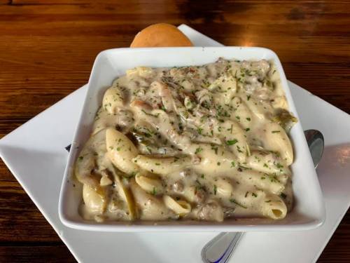 A bowl of creamy pasta with herbs, featuring macaroni and a side roll on a wooden table.