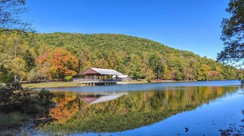 A serene lake scene with a wooden pavilion, surrounded by colorful autumn trees and a clear blue sky.