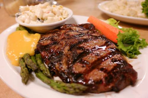 A grilled steak served with asparagus, cheese sauce, a carrot, and a side of macaroni salad on a white plate.