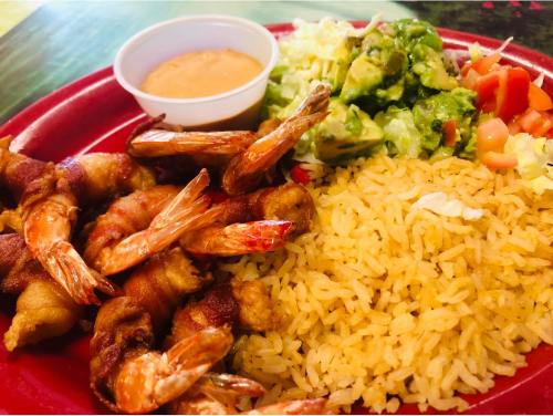 A red plate with shrimp wrapped in bacon, rice, salad with avocado, and a small cup of sauce.