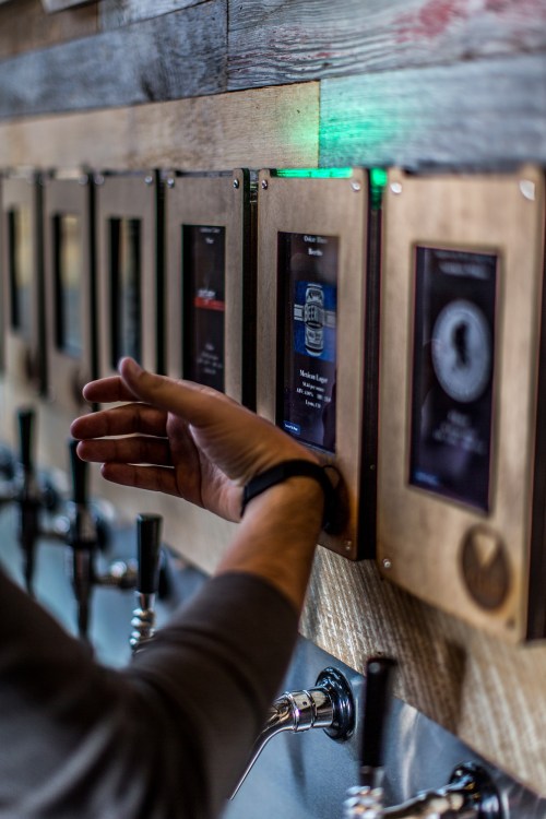 A person interacts with a digital beer tap display, selecting a drink from a wall-mounted menu.