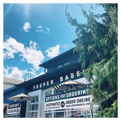 A storefront with the sign "Proper Bagel," featuring outdoor seating and a clear blue sky above.