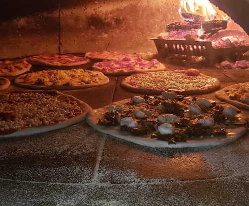 Pizzas baking in a wood-fired oven, with flames and various toppings visible on the crusts.